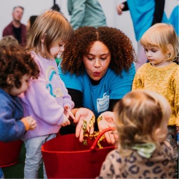 Travelling Light facilitator is blowing gold confetti into a red bucket with young children standing around her watching. Photo by Alastair Brookes.