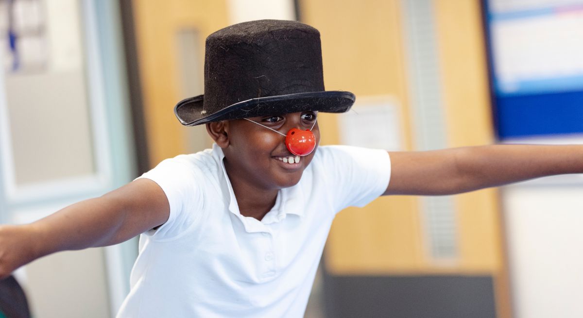 A photo of a boy wearing a red nose and a black top hat, white t-shirt and his arms stretched out horizontally to either side of him.