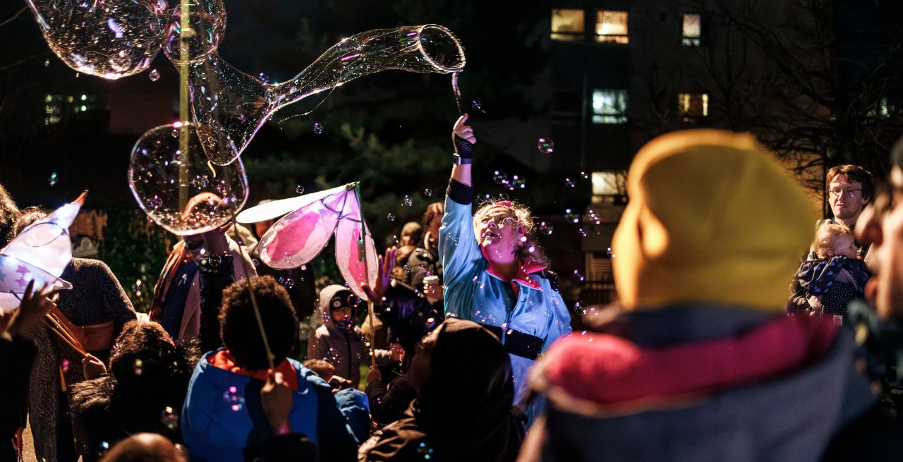 Youth theatre members face inwards in a circle with arms raised together - performing at Winter Lights with audience members looking on. 