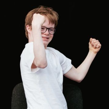 A member of Louder Than Words has both arms raised with fists clenched. They are white, with short hair, wearing a white t-shirt and and black rimmed glasses.