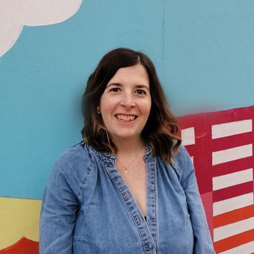Lizzy Stephens - a photo of Lizzy standing outside of Travelling Light looking towards the camera. The wall behind is painted with a mural of Barton Hill. Blue sky and a red building.