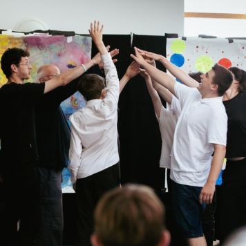 Louder Than Words members and facilitators performing during their sharing in 2022. The group stands in a circle with their arms raised and hands coming together in the middle of circle. 