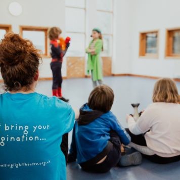 An image of a Youth Theatre member taking part in a session. They are white with mid-length dark hair, wearing a blue jumper and brown trousers, kneeling on one knee with arms outstretched to their sides.