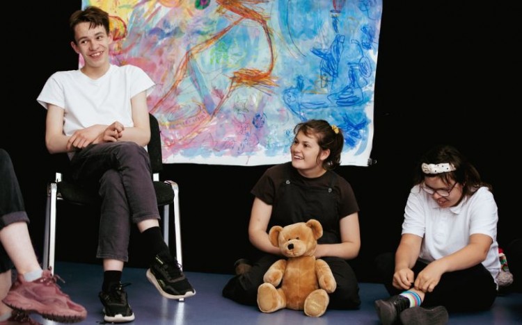 Louder Than Words members and facilitators performing during their sharing in 2022. A member sits on a chair, while 2 members sit on the floor, with a colourful painting behind them.