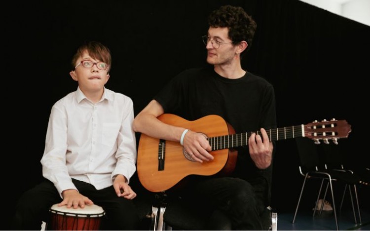 A member of Louder Than Words sits and plays the drum while Louder Than Words assistant Bruno sits next to him playing the guitar.