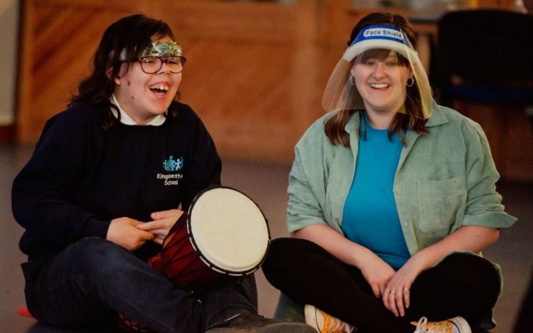 A member of Louder Than Words sits cross legged on the floor on the left of photo with a hand drum between her legs. Amy, one of our Louder Than Words assistant facilitators sits on the right.